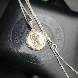 May include: Silver chain necklace with a round pendant featuring a depiction of Saint Christopher and the text "Saint Christopher Protector".