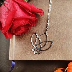 以下が含まれることがあります： 茶色の革張りの本の上に飾られた、花の模様のシルバーペンダントネックレス。鮮やかな赤いバラとオレンジ色のホオズキの花も画像にあります。ネックレスは繊細なチェーンから吊り下げられています。