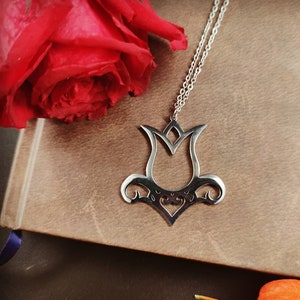Peut inclure: Un collier pendentif en argent avec un motif de tulipe stylisé. Le pendentif est suspendu à une chaîne délicate et repose sur un carnet en cuir marron. Une rose rouge et des baies oranges sont en arrière-plan.