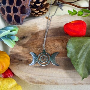 May include: A silver necklace with a triple moon pendant and a Celtic knot design. The pendant hangs from a fine chain. The necklace is presented on a wooden surface, surrounded by decorative items such as a red flower, green leaves, and small pumpkins.