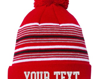 May include: Red knit beanie with a white pom pom and black and white stripes. The beanie has the text "YOUR TEXT" embroidered on the front in white.
