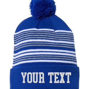 Puede incluir: Gorro de punto azul y blanco a rayas con un pompón azul en la parte superior. El gorro tiene un puño abatible con el texto "YOUR TEXT" en blanco.