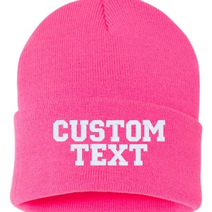 May include: A bright pink beanie with white text that reads "CUSTOM TEXT".
