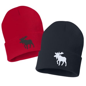 Op de afbeelding: Twee gebreide mutsen, een rode en een donkerblauwe, met een geborduurd elandsilhouet aan de voorkant. De eland op de rode muts is zwart, en de eland op de donkerblauwe muts is wit.