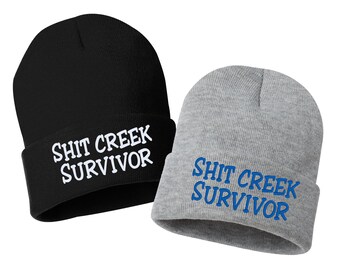 May include: Two knit beanies with the text "Shit Creek Survivor" embroidered on them. One beanie is black with white text and the other is gray with blue text.
