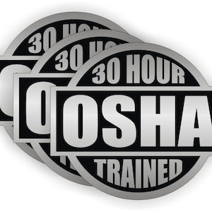Op de afbeelding: Een stapel zilveren en zwarte OSHA-gecertificeerde stickers. Elke sticker heeft een cirkelvormige rand met de woorden "30 HOUR" bovenaan en "TRAINED" onderaan. Het woord "OSHA" staat in grote witte letters in het midden.