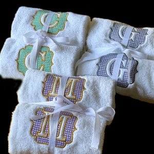 May include: White terry cloth towels with embroidered monogram letters in teal, purple, and gray, each tied with a white ribbon. The towels are folded and stacked, showcasing the decorative monogrammed designs.