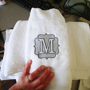 May include: White terry cloth hand towel with an embroidered monogram letter "M" in a decorative frame. The towel is folded and stacked with other towels. A hand with a ring is holding the towel.