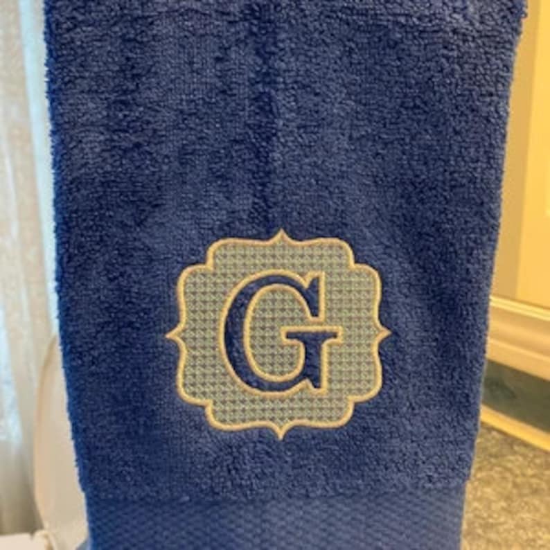 May include: A navy blue hand towel with an embroidered monogram letter "G" in a decorative frame. The frame has a light blue and gold pattern.