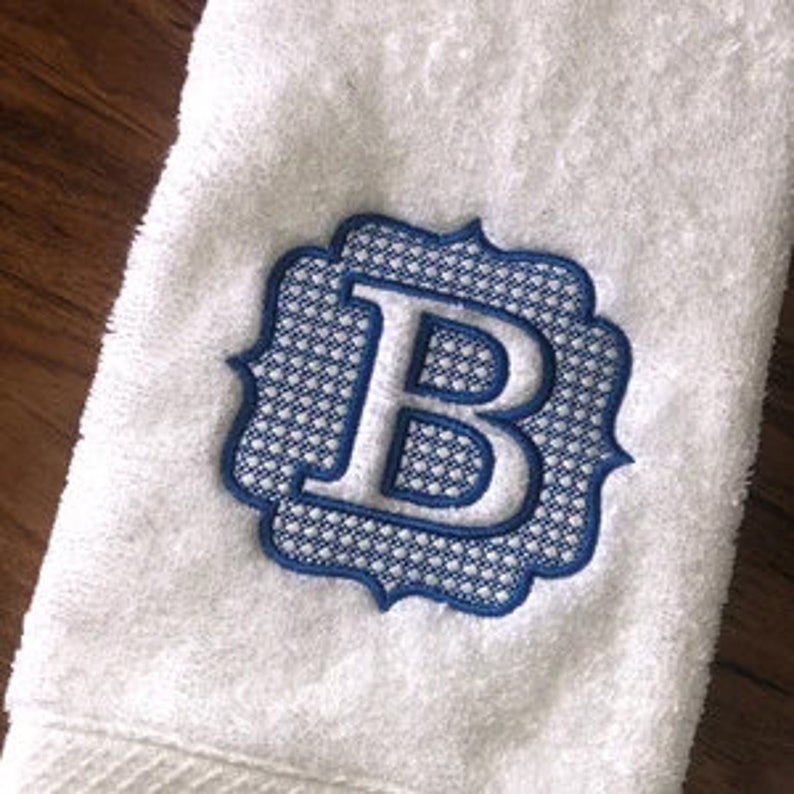 May include: White towel with a blue embroidered letter "B" in a decorative frame. The embroidery has a detailed, textured appearance, and the towel is folded.