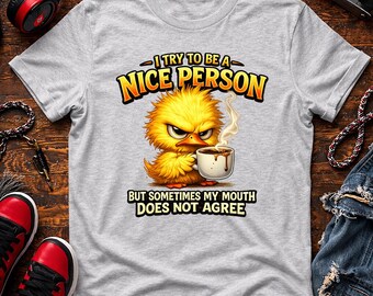 May include: A light gray t-shirt featuring a cartoon yellow chick with an angry expression, holding a coffee cup. The shirt has the text "I TRY TO BE A NICE PERSON" above the chick and "BUT SOMETIMES MY MOUTH DOES NOT AGREE" below.