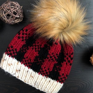 May include: A red and black plaid knit beanie with a white cuff and a brown faux fur pom pom.