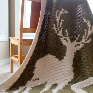 May include: A crocheted blanket with a white deer silhouette on a green background. The blanket is folded over a wooden chair.