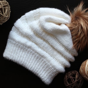 May include: White knitted beanie with a brown faux fur pom pom. The beanie is folded over and has a slouchy look.