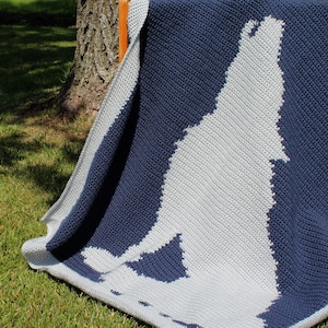 Puede incluir: Una manta de crochet con una silueta de lobo blanca sobre un fondo azul marino. La manta está doblada y drapeada sobre una silla de madera.