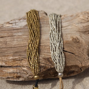 以下が含まれることがあります： 金と銀の2本の編み込みブレスレットが、流木の上に置かれています。ブレスレットは、小さな正方形のビーズで作られています。