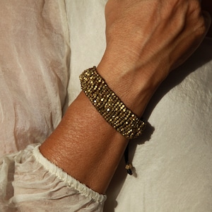 Könnte beinhalten: Ein goldfarbenes Armband mit einem schwarzen Kordelverschluss. Das Armband besteht aus kleinen, runden Perlen, die eng aneinander gewebt sind.
