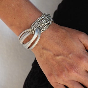 Könnte beinhalten: Ein silbernes Armband mit mehreren Reihen kleiner Perlen und einer Lederumwicklung. Die Lederumwicklung ist silberfarben und hat ein geknotetes Design.