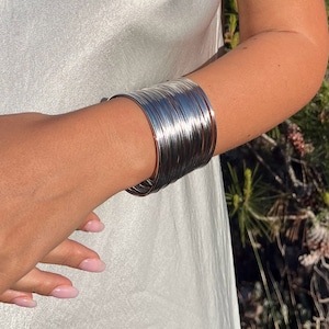 May include: A silver bangle bracelet made of multiple thin metal bands stacked on top of each other. The bracelet is worn on a person's wrist.