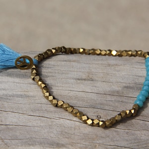 Puede incluir: Un brazalete de cuentas doradas y azules con un dije de signo de paz y una borla azul.