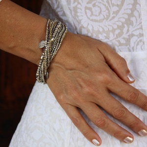 Op de afbeelding: Een close-up van iemands pols met een zilveren en gouden kralen armband met een decoratieve hanger. De armband is gelaagd en heeft een bohemian stijl.