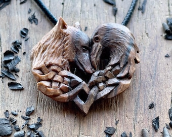 May include: A hand-carved wooden pendant featuring the profiles of a wolf and a raven facing each other, their forms intertwined in a heart-shaped design. The pendant is suspended from a thin black cord and displays detailed wood grain textures.