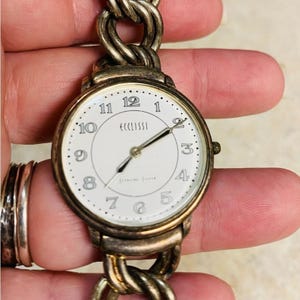 May include: A vintage Eccissi wristwatch with a white face, black numerals, and gold-tone hands. The watch has a gold-tone bezel and is attached to a chain-link bracelet. The watch is a classic accessory.