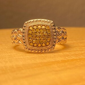 May include: A gold-toned ring with a square, yellow gemstone cluster center. The ring has a braided band and a detailed, textured setting around the gemstones. The ring is displayed on a wooden surface.