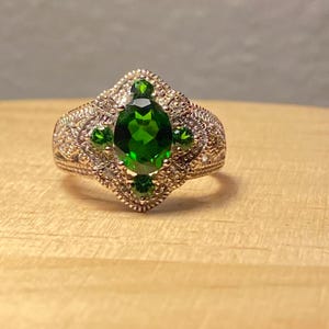 May include: A gold-toned ring with an oval, emerald-green gemstone at the center, flanked by smaller green stones. The band features intricate filigree detailing and small, clear accent stones, creating a vintage aesthetic.