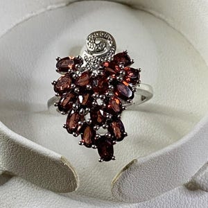 May include: A silver ring featuring a cluster of oval-cut, deep red gemstones, resembling a bunch of grapes. The ring has a decorative swirl design at the top and is set against a white background. A unique piece of jewelry.