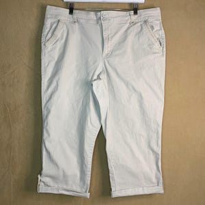 Könnte beinhalten: Hellbeige Caprihose mit Knopfverschluss, Vordertaschen und umgeschlagenem Saum. Die Hose hängt an einem weißen Kleiderbügel vor neutralem Hintergrund. Die Caprihose ist aus leichtem Stoff gefertigt.