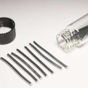 Könnte beinhalten: Eine kleine Glasflasche gefüllt mit silberfarbenem Draht, mit einem schwarzen Plastikdeckel und mehreren Drahtstücken, die auf einer weißen Oberfläche liegen.