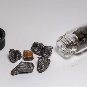 Op de afbeelding: Een klein glazen flesje gevuld met grijze metalen stukjes van het element osmium. Een paar grotere stukjes osmium liggen verspreid op een wit oppervlak naast het flesje. Het flesje heeft een zwarte plastic dop.