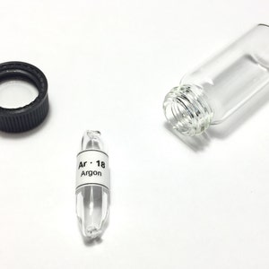 Puede incluir: Un vial de vidrio transparente con una tapa negra y una botella de vidrio separada. También hay un pequeño vial etiquetado "Ar · 18 Argon". Los elementos están sobre una superficie blanca.
