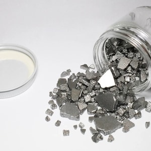 Op de afbeelding: Een glazen pot met een wit deksel, gevuld met kleine, onregelmatig gevormde stukjes zilvergrijs metaal. De pot is omgevallen en laat de metaalstukjes op een wit oppervlak vallen.