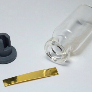 Könnte beinhalten: Eine klare Glasflasche mit einem grauen Gummistopfen und einem dünnen goldenen Streifen. Die Flasche ist leer und der Stopfen ist so konzipiert, dass er fest sitzt.