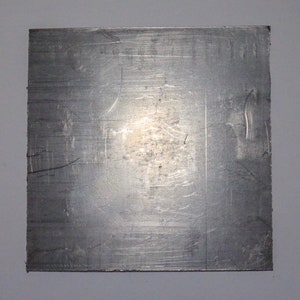 May include: A square, metallic sheet with a brushed, silver finish. The surface has visible scratches and imperfections, creating a textured appearance. The edges are clean and straight, suggesting a manufactured product.