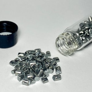 Op de afbeelding: Een klein glazen flesje gevuld met zilverkleurige metalen stukjes, met een zwarte plastic dop en enkele metalen stukjes die op een wit oppervlak zijn gemorst.