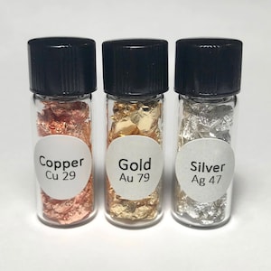 May include: Three small glass bottles with black lids containing flakes of copper, gold, and silver. Each bottle has a white label with the name of the metal and its atomic number.