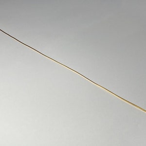 Puede incluir: Un alambre delgado y recto, de color dorado, probablemente para la fabricación de joyas o manualidades, está posicionado en diagonal sobre un fondo gris claro liso. El alambre parece ser de metal y tiene una superficie lisa y reflectante.