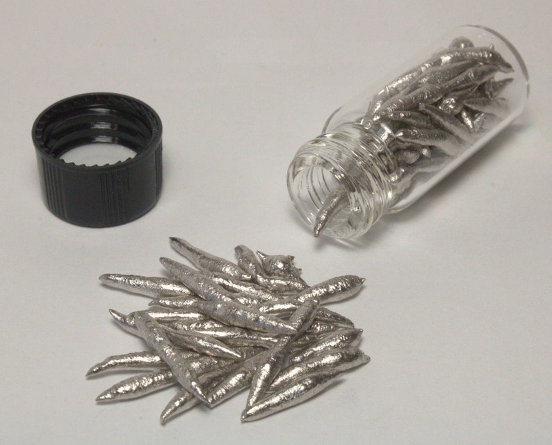 Op de afbeelding: Een glazen flesje gevuld met kleine, zilverkleurige metalen stukjes. Het flesje ligt op zijn zijkant en enkele stukjes zijn op een wit oppervlak gevallen.