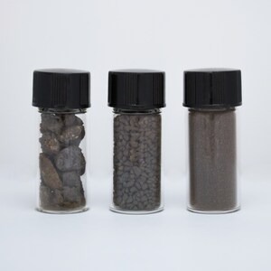 Puede incluir: Tres pequeños viales de vidrio con tapas de rosca negras, cada uno contiene un tipo diferente de arena o roca gris oscuro.