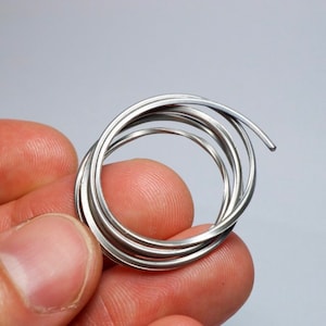 Op de afbeelding: Een opgerold stuk zilverkleurige metaaldraad gevormd in een cirkelvorm met meerdere overlappende lussen. De draad heeft een glad, reflecterend oppervlak en een consistente dikte over de gehele lengte, wat een eenvoudige spiraalstructuur creëert.