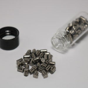 May include: A small glass bottle filled with silver metal pellets. The bottle is open and some of the pellets have spilled out onto a white surface.