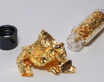 May include: A small glass bottle filled with thin, gold-coloured flakes. The bottle is open and some of the flakes are scattered on a white surface.