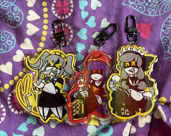 May include: Three acrylic keychains featuring anime-style characters with black metal clasps. The characters have yellow, red, and gray color schemes. The keychains are on a purple patterned background.
