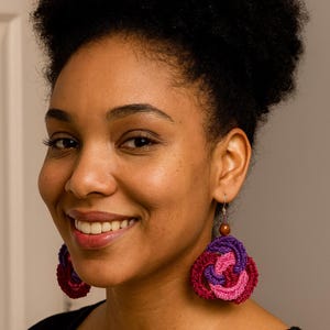 May include: Close-up of a person wearing large, handmade crochet earrings. The earrings feature a spiral design with shades of pink, purple, and burgundy. A small wooden bead tops each earring, suspended from a silver hook.