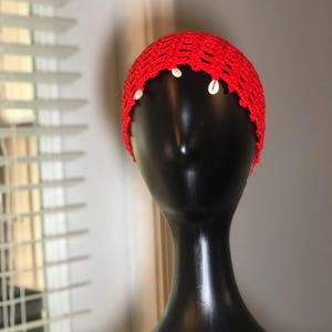 May include: A vibrant red crochet head covering, adorned with small white seashells along the scalloped edge. The head covering is displayed on a black mannequin head, showcasing its texture and design.