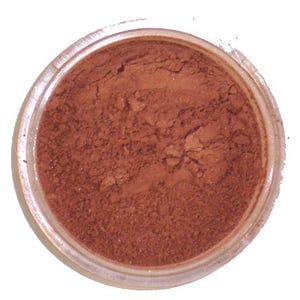 Può includere: Un piccolo barattolo trasparente pieno di polvere marrone-rossastra. La polvere ha un aspetto fine e strutturato, e il barattolo è posto su uno sfondo bianco. Il prodotto sembra essere un cosmetico, possibilmente ombretto o fard.