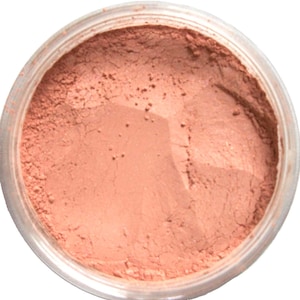 Può includere: Un contenitore rotondo e trasparente riempito di polvere color pesca. La polvere ha un aspetto fine e strutturato e il contenitore è visto dall'alto. Il prodotto sembra essere un cosmetico, come un fard o un ombretto.